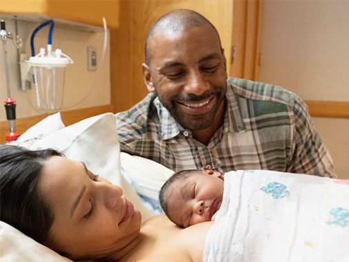 husband admiring his recovering wife with their newborn baby on her chest